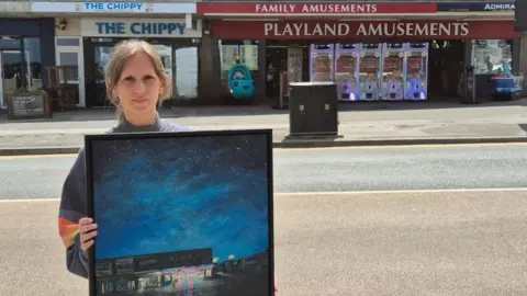 A picture of Dianne holding a picture of her blue painting outside of the arcade. 