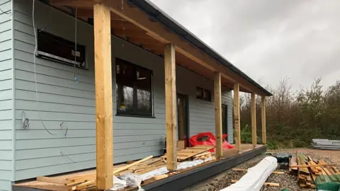 A wooden building painted light blue with a veranda outside is still under construction. All the windows and doors are fitted but there are a quantity of timbers lying around the site and on the veranda 