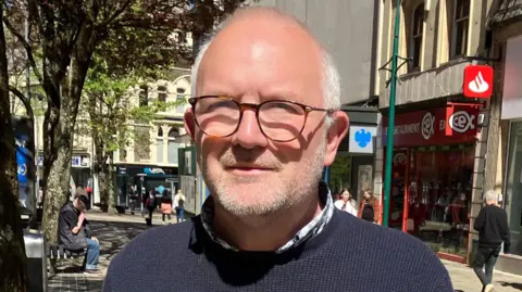 Andrew Lightbown is wearing a dark blue jumper and is looking at the camera. His arms and upper body are visible. People can be seen walking in the background in front of the city's shops.