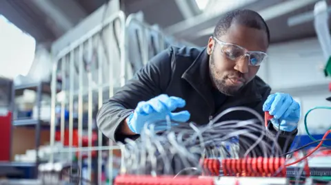 Getty Images Man wearing safety goggles and blue gloves looking over some wires on a table in a manufacturing plant