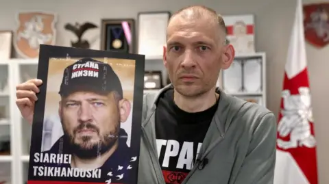 Francesco Tosto/BBC Sergei Tikhanovsky holds a poster of himself before being arrested. It shows him in good health, wearing a black baseball hat and bearded, in contract to the gaunt, clean-shaven image of him after the release