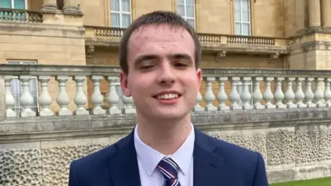A young man wearing a navy jacket, white shirt and striped blue, white and red tie. He has short brown hair and is smiling. In the background is a grand looking building, with columns. 