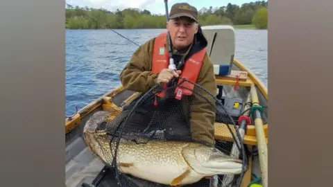 Submitted A man wearing a red life jacket and a khaki jacket is posing on a boat in the middle of a lake with a giant pike in a fishing net.