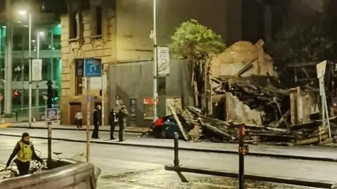 A pile of rubble on top of a car on a street at nighttime while three people look on an a police officer holds a bike in the foreground