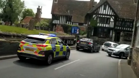 TAG A262 A police car with flashing sirens sits behind traffic stuck at a narrow corner