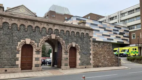 The entrance to Jersey General Hospital on a grey day. There is an ornate gatehouse with three arched gateways and to the right inside the premises are two parked ambulances. There is a road lined with a pavement in the foreground.