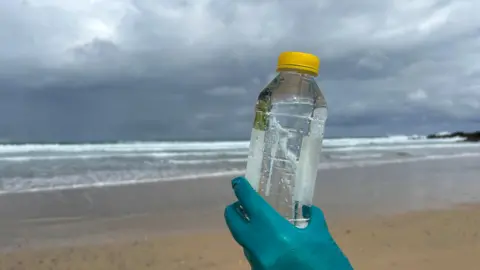 Environment Agency A hand - wearing a protective glove that is green - is holding up a plastic bottle containing water at a beach. It is a sample of the bathing water, there is golden sand and breaking waves in the background. The sky is cloudy. 