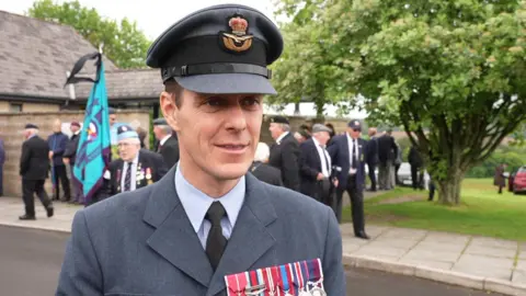 Gareth Elliot looking to the right and smiling. He has a row of medals on his jacket and is stood in front of a building, with people stood outside of it. 