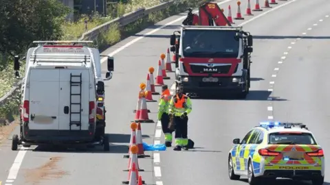 A close-up view of a motorway lane shows orange traffic cones stretching into the distance. A marked police car with blue lights, a white recovery van and a red recovery lorry are parked in the closed lanes. Two highway workers wearing hi-vis clothing stand beside the cones.