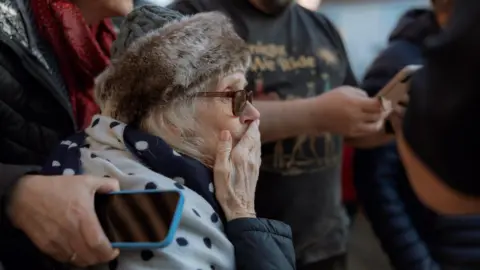 Jessica Moore Photography Sam King's mother, pictured side-on, is looking to the right of the frame sitting amongst a crowd of people. She has her hand over her mouth. She has glasses and thick winter clothing on, including a fur hat.