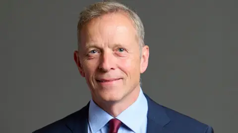 An official portrait of Matt Rodda, who is wearing a dark blue jacket, light blue shirt and maroon tie 