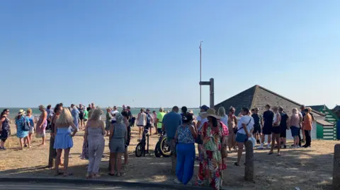 People gathering on a beach. It is a sunny evening. The sea is in the distance and roofs of huts can be seen.
