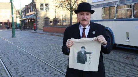 Beamish Ian Bean is wearing a historical costume consisting of a bowler hat and a smart black suit. He is holding up a tote bag with his own image on. The text next to him reads: "SLAY".