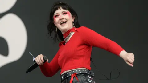 Getty Images A woman performing on stage. She has black hair and is wearing red face paint and a red jumper. He is holding a microphone. She looks like she is dancing and is smiling.