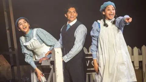 James Glossop Three Railway Children actors stand on a platform and look to their left. One is pointing and all are smiling.
