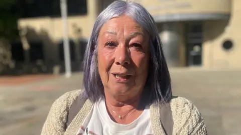 A woman with silvery purple hair looks directly at the camera outside a court building.