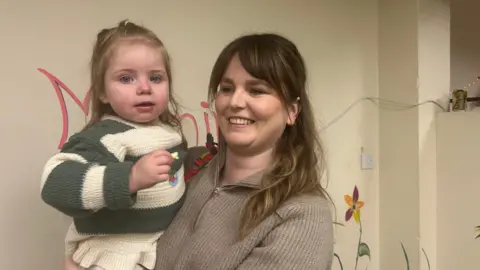 Emma Gould holding her daughter. She is smiling and wearing a grey jumper. Her daughter is wearing a white and green striped jumper. 