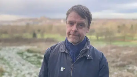 A man standing in a field. He has short brown hair and brown/grey stubble. He is wearing a navy jacket with a white flower pin badge. He has a blue scarf.