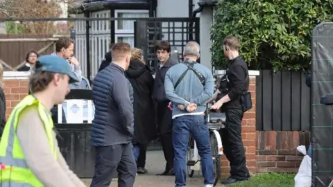 Paul Mescal is dressed in a blue jacket black trousers on a film set outside a beige coloured semi detached house.