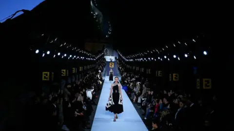 Getty Images models wear black and white clothes walking in a long running run by the lights, oh the Great Wall of China, surrounded by people sitting and watching the fashion show