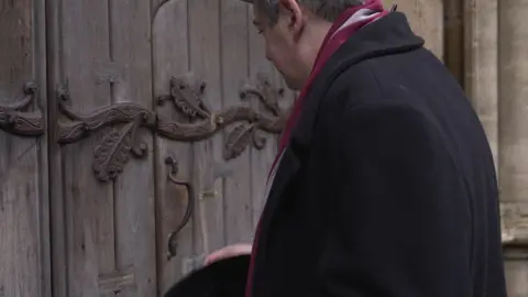 A man wearing a long, black coat has removed his hat before entering an old, ornate door.