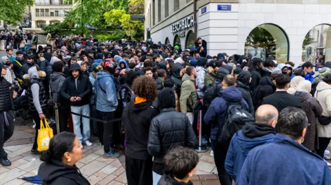 EPA/Shutterstock A large crowd of people stand around outside a building