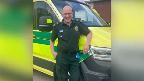 GRAHAM MACMILLAN Graham McMillan wearing a dark green ambulance uniform with a blue NHS badge. He is smiling at the camera and leaning on an ambulance.