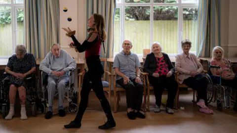 PA Media A woman juggling three multi-coloured balls in front of an audience of care home residents.