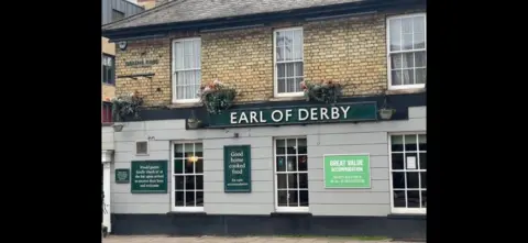 Robbie Kalus/BBC The Earl of Derby pub: a brown and grey building with white-framed windows. The words "Earl of Derby" are written in white capital letters on a green sign fixed to the front of the pub. Below the sign are signs advertising "good home cooked food" and "great value accommodation".