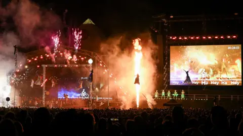 Reuters/ Jaimi Joy Acrobats perform at the Pyramid arena during Glastonbury Festival, there is a large screen to the right of the stage and bright sparks around the stage.