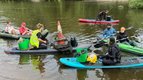 Seven people on paddleboards and small inflatable boats paddling on a river, with a collection of objects piled up on them, including two red traffic cones