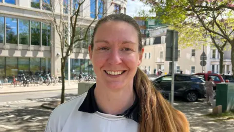 Woman with long dark hair tied back looking into the camera and smiling. She is wearing a white jumper and you can see the collar of a black top underneath it. There are a couple of trees behind her, a pavement, a road and a tall building in the background with bikes outside it. The sun is shining.
