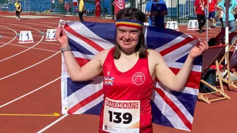 A woman smiling at the camera, hands aloft holding a union jack flag behind her back. wearing a red athletic vest and standing on an athletics track. She's also wearing a rainbow coloured sweat band on her head.