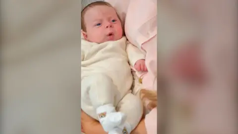Baby wearing yellow onesie being held by woman in pink shirt