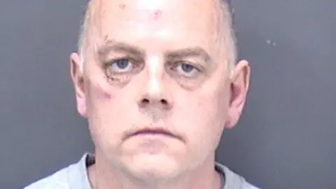 In a police custody photo, Paul Hopkins appears to have bruising around his right eye and a mark on his forehead. He has short grey hair and wears a grey, collarless top.
