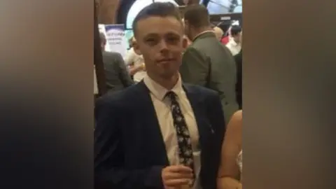 Family handout Kailem Gibson has short hair, a dark jacket, a white shirt and black patterned tie and is holding a drink.