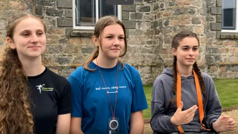 Three girls are standing outside in front of a stone building. They are all aged 13 or 14 and have long hair. One girl has a black T-shirt, whilst another is in a blue T-short and the girl on the right of the image who has long plaits has a grey hoodie.