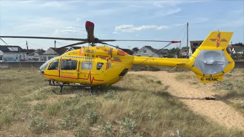 A yellow air ambulance helicopter sitting on a beach in Jaywick. It is branded as the East Anglian Air Ambulance. The rotor blades are not turning, and there is a grey covering over the tail rotor.