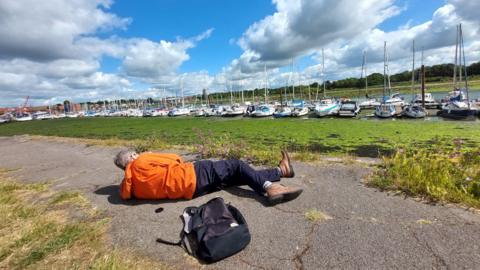 THURSDAY - A man in an orange jacket lies down on a path taking photos of yachts in the water in the distance om Fareham