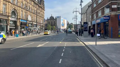 A road showing part of it with a police cordon blocking off access. On each side there are buildings and several pedestrians. There are two police cars on the road.