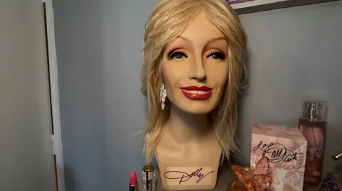 The image is a close-up of a display featuring a mannequin-style head mounted on a stand and fitted with a blonde wig. 