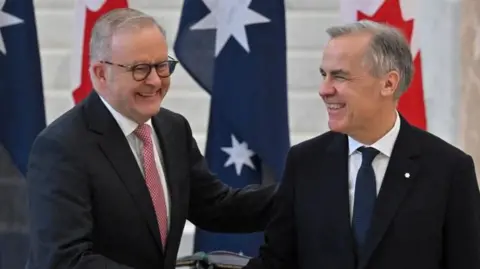 Two middle aged men with grey hair and wearing dark suits smile in front of the Australian and Canadian flags.