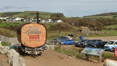 Google A pine-coloured wood sauna on a stony beach with a black chimney. The structure is on wheels. In the background there are several cars. On an elevated piece of ground behind the sauna, there are several caravans.