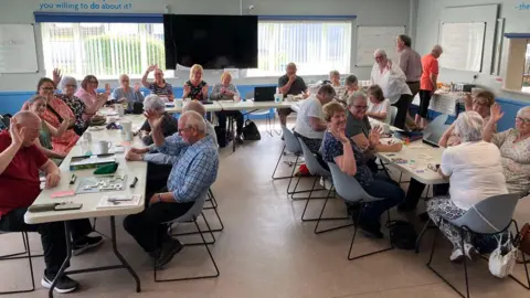 The Centre West Cumbria Dozens of people are sitting around tables at a Biccies and Banter session. Many are waving at the camera. Several are playing games such as Scrabble.