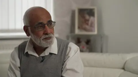 Man with grey beard in grey jumper and white shirt on sofa. He is wearing glasses
