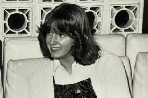Lynne Darcy An old black and white picture of Lynne Darcy. She is around 14 years ols and has shoulder length hair which is styled and flicked back from her face. She is wearing a white cardigan over a white blouse and darker coloured top. 
