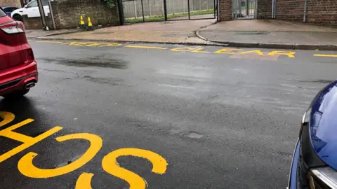 The image shows repainted yellow zigzags and 'School Keep Clear' warnings outside the former site of Halfway Houses Primary School in Sheerness, Kent. A small section of a red car can be seen in the image, and a small section of a separate blue car can also be seen. 
