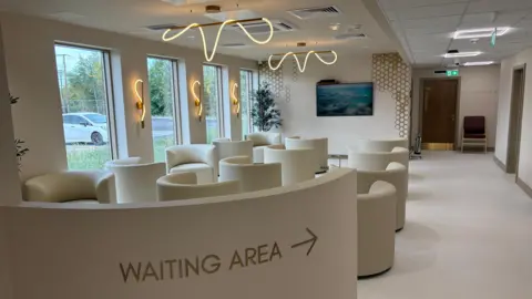 A waiting are with beige walls, curved soft seats and long, floor-to-ceiling windows. Contemporary light fittings on the wall and ceiling throw a warm light across the room. There is television on the back wall, and a door to the right.
