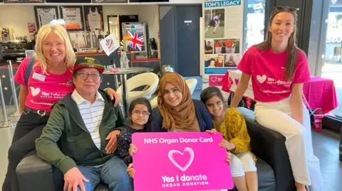 From left to right, Lisa Wilson with recipient Gordon, recipient Fatima and her mother Lubna and sister Sofia, and her daughter Pippa. They are sitting on a sofa, smiling for the camera. The mother and two daughters are holding a pink NHS Organ Donor Card with a heart in the middle and the message "Yes I donate". Various Transplant Games merchandise is displayed behind them, as well as information about Lisa's son, Tom.