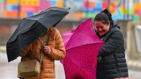 PA Media Two women struggle with umbrellas in the rain in Liverpool